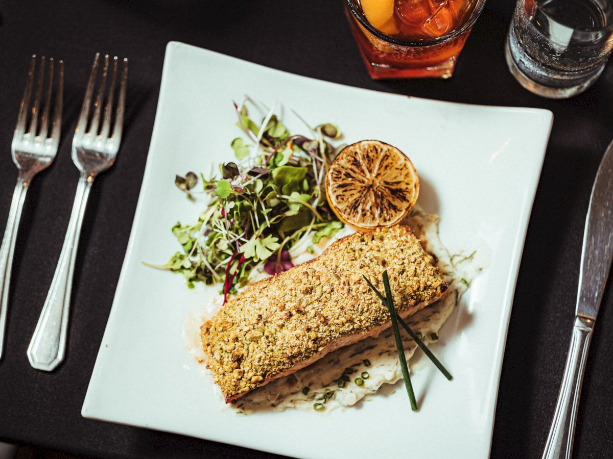 A gourmet dish featuring a breaded fillet with mixed greens, a lemon slice, and chives on a white plate, plus an iced drink and cutlery.