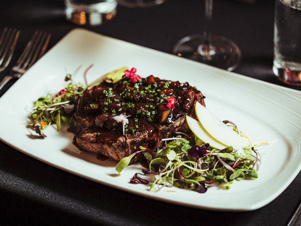 A gourmet dish featuring a piece of meat topped with herbs, alongside fresh greens, garnished with flowers and apple slices, on a white plate.