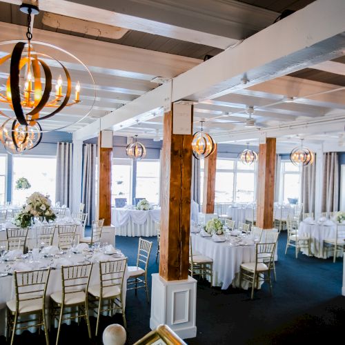 A beautifully decorated event space with round and rectangular tables set for a formal occasion, featuring elegant lighting fixtures and floral centerpieces.