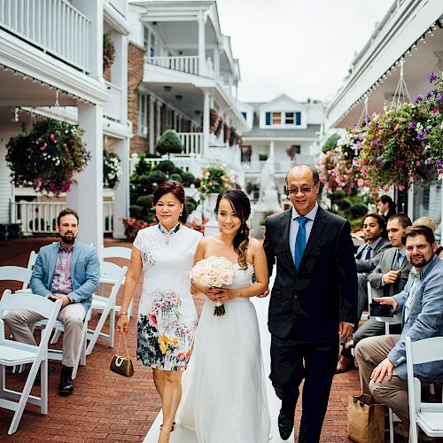 A bride is walking down the aisle with two people, surrounded by seated guests in an outdoor wedding setting with hanging flower baskets and white chairs.