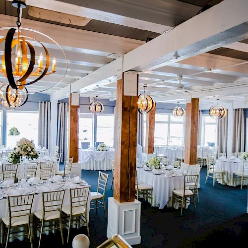 An elegantly decorated dining room with round and rectangular tables, white chairs, floral centerpieces, and modern chandeliers hanging from the ceiling.