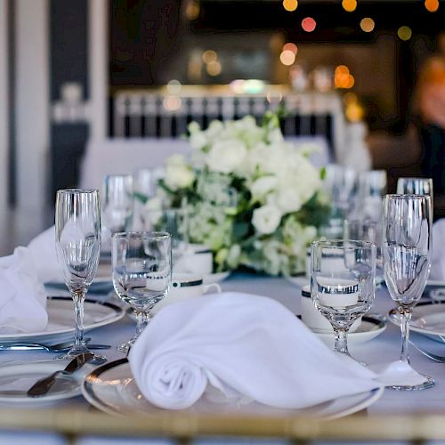 A beautifully set table with white napkins, plates, glasses, and a central floral arrangement of white flowers in the background.