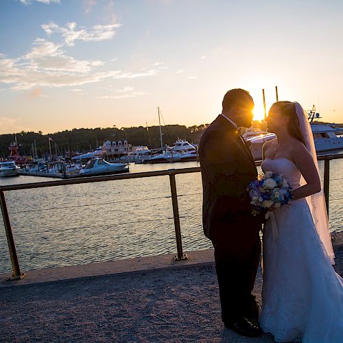 A bride and groom share a romantic moment by the waterfront at sunset, with boats and a marina in the background.