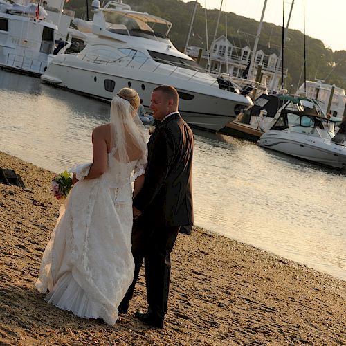 A bride and groom stand on a sandy shore by a marina, holding hands, with boats and yachts docked in the calm water.