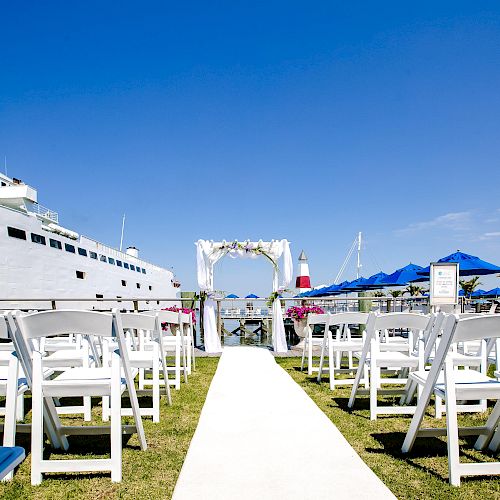 An outdoor ceremony setup with white chairs on either side of a white aisle, facing an archway, with a large boat and blue sky in the background.