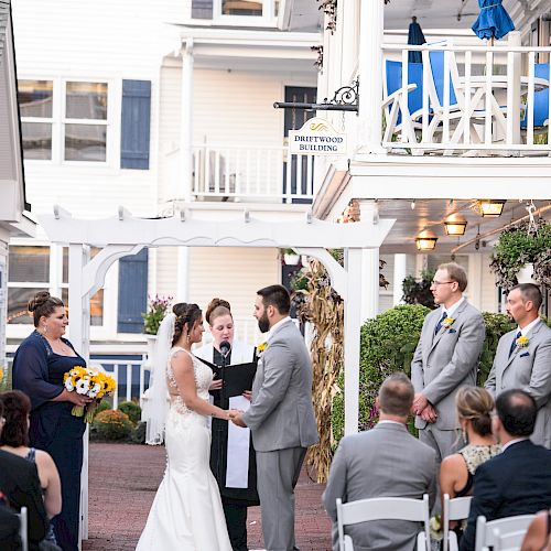 A wedding ceremony is taking place outdoors with a bride and groom at the altar surrounded by their wedding party and guests in an intimate setting.