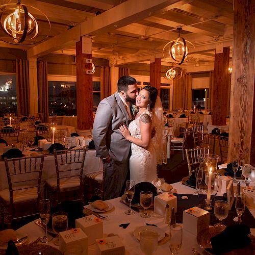 A couple dressed in wedding attire embraces in a beautifully decorated reception venue with tables set for dining and elegant lighting fixtures.