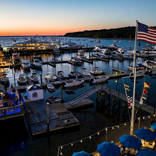 A marina at sunset with numerous boats docked, an American flag in the foreground, and outdoor seating area with blue umbrellas lit up.