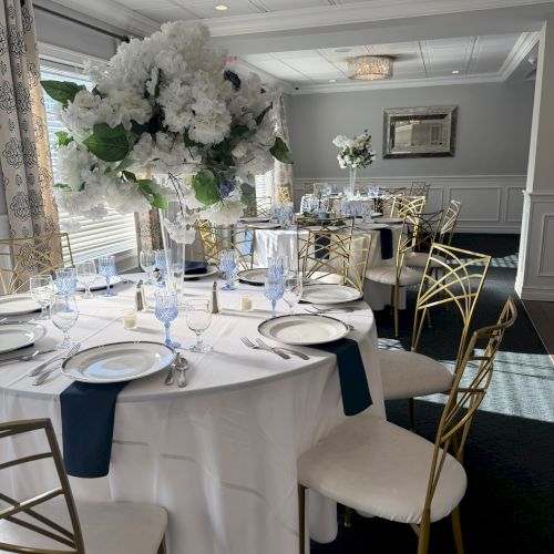 A formal banquet setup with round tables, white tablecloths, tall white floral centerpieces, and gold chairs in an elegant, softly lit room.
