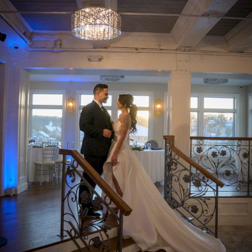 A couple stands on a lit staircase, the bride in a white wedding gown and veil, the groom in dark attire, posing indoors with blue lighting.