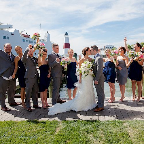 A wedding party celebrates near a waterfront with a ship and lighthouse in the background, while the bride and groom kiss in the center.