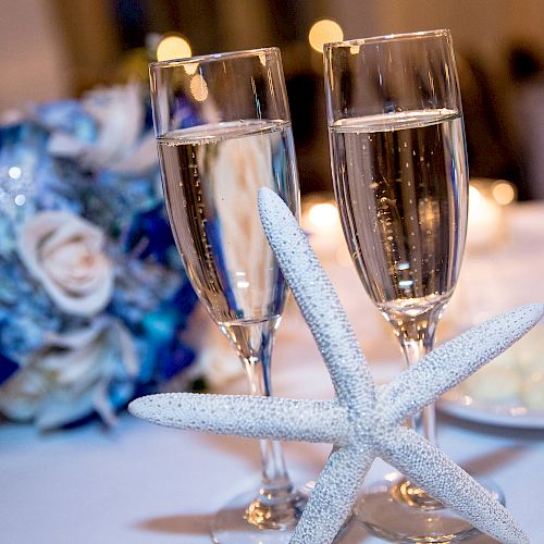 Two champagne glasses with a starfish decoration and a bouquet of flowers in the background on a table.