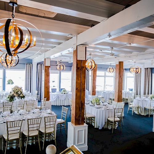 The image shows a beautifully decorated event space with round tables, chairs, floral centerpieces, and modern light fixtures, ready for a formal gathering.