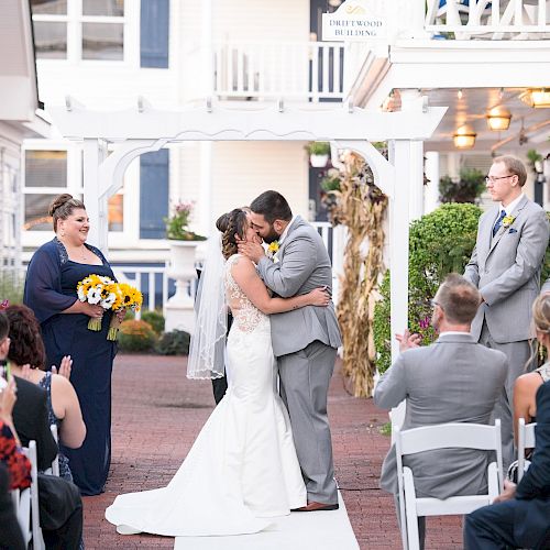 A couple shares a kiss at an outdoor wedding ceremony, with the bridal party and guests celebrating around them.