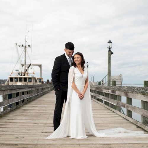 A newlywed couple stands on a wooden pier, the groom in a black suit and the bride in a white gown, with a lighthouse in the background.