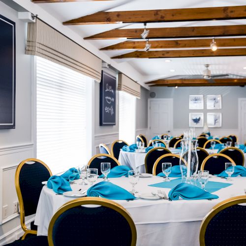 A bright dining room with round tables, blue napkins, black-and-gold chairs, white walls, and chalkboard artwork along the left wall.