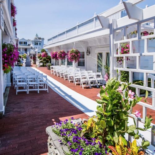 A sunny seaside terrace with white chairs lined up, flower pots, and a brick walkway, perfect for outdoor dining by the water.