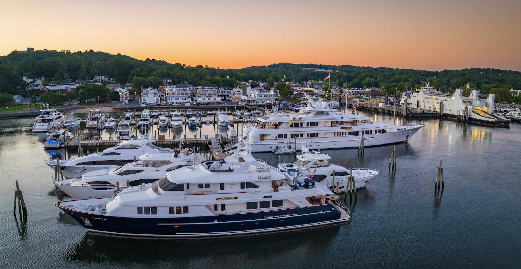 The image shows a marina filled with yachts and boats, nestled against a backdrop of a coastal town at sunset.