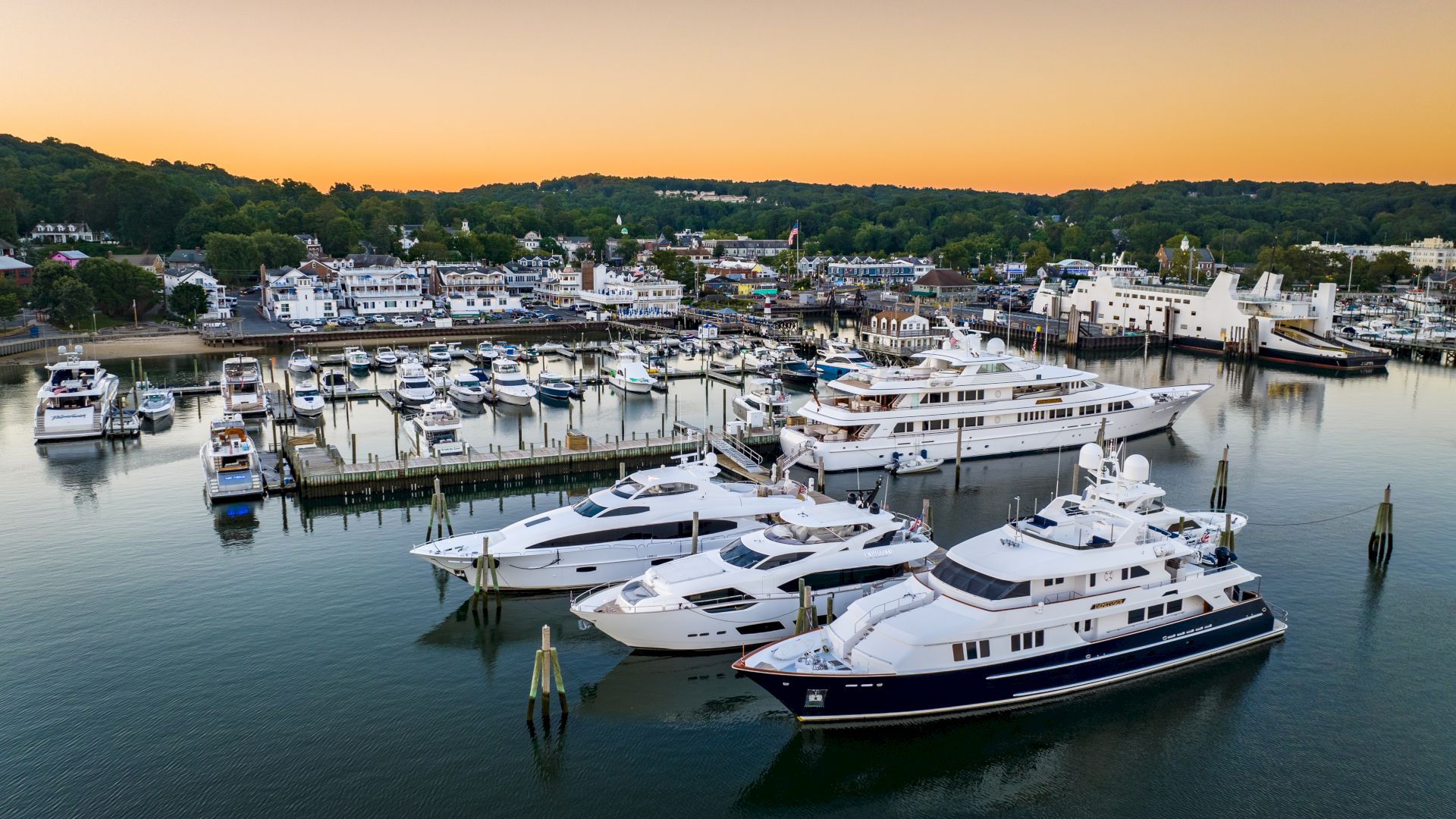 The image shows a marina with several luxury yachts docked, surrounded by calm water and a backdrop of buildings and trees at sunset.