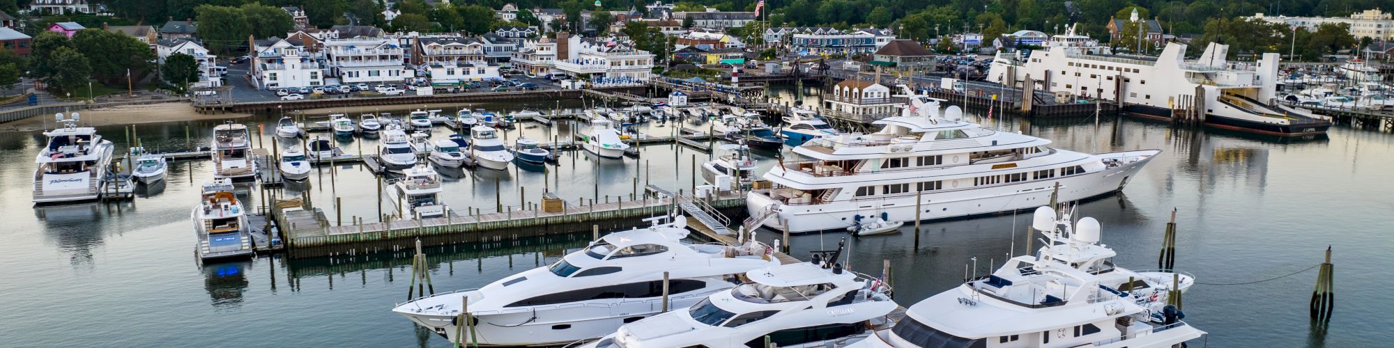 The image shows a marina with several luxury yachts docked, surrounded by calm water and a backdrop of buildings and trees at sunset.