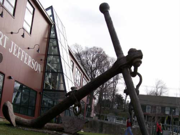 The image shows a large anchor displayed outside a building with "PORT JEFFERSON" written on it and a glass atrium. Two people are visible in the background.