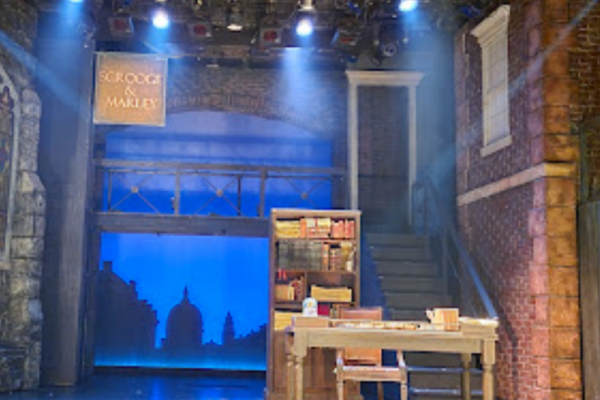 The image shows a theater stage set with a desk, bookshelves, stairs, and a sign that says 