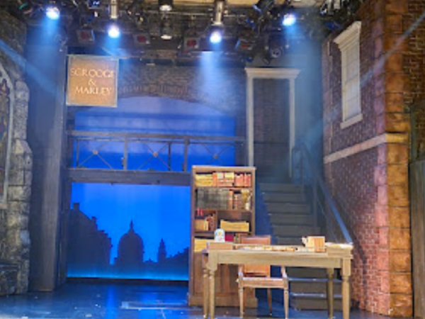 The image shows a theater stage set with a desk, bookshelves, stairs, and a sign that says "Scrooge and Marley," likely for a play adaptation of "A Christmas Carol."