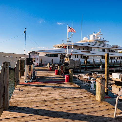 A large luxury yacht docked at a wooden pier with various equipment and boats, under a clear blue sky, and an American flag flying on the yacht.