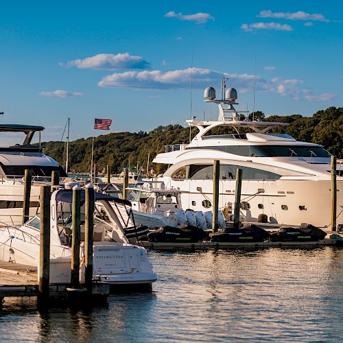 The image shows a marina with several yachts docked, a clear blue sky, and trees in the background, with the American flag visible.