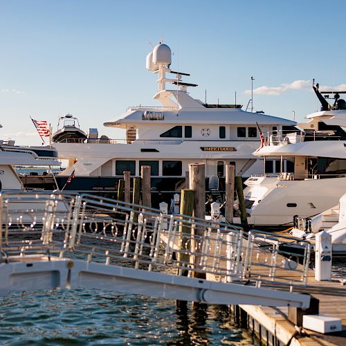 The image shows a marina with several large yachts docked, connected by walkways under a clear blue sky, and some flags are visible.