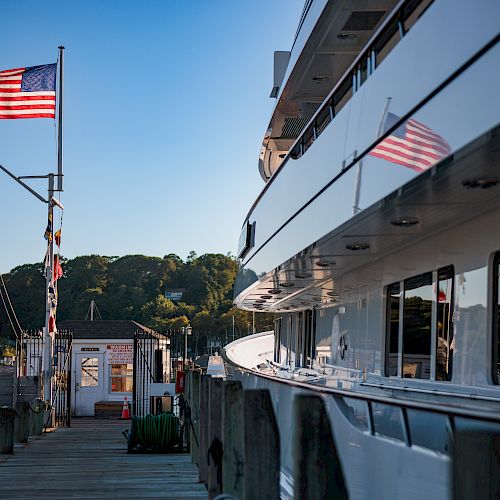 A dock with an American flag, a small building, and a large boat reflected in the water. Trees and a clear blue sky are in the background.