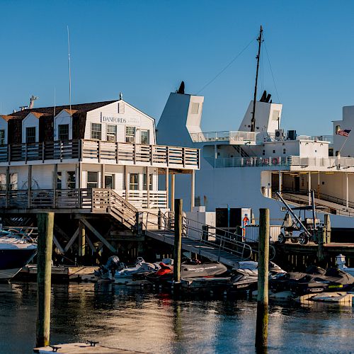 A marina with boats docked and a two-story building next to a large ship in sunny weather. Several jet skis are also visible in the water.