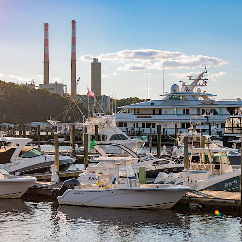 Several boats and yachts are docked at a marina with industrial buildings and smokestacks in the background, under a partly cloudy sky.