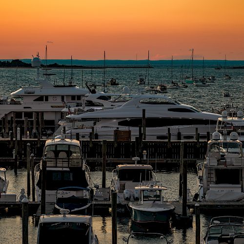 The image shows a marina filled with various yachts and boats during sunset, with a calm sea and the horizon visible in the background.