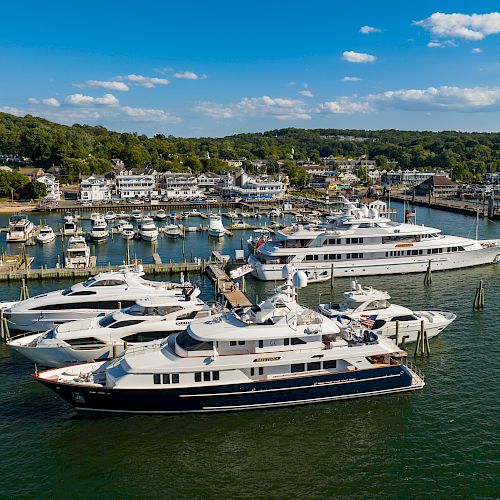 The image shows a marina filled with various yachts and boats docked, with a coastal town and green hills in the background under a blue, partly cloudy sky.