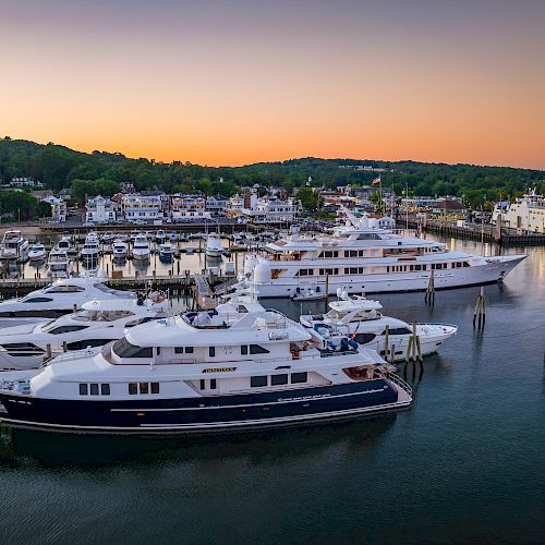 The image shows a marina with several luxury yachts docked, set against a calm waterway and a sunset-lit landscape with buildings and trees in the background.