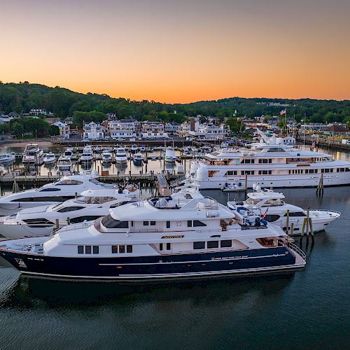 A marina with several yachts docked, set against a scenic backdrop with a sunset sky and greenery in the distance.