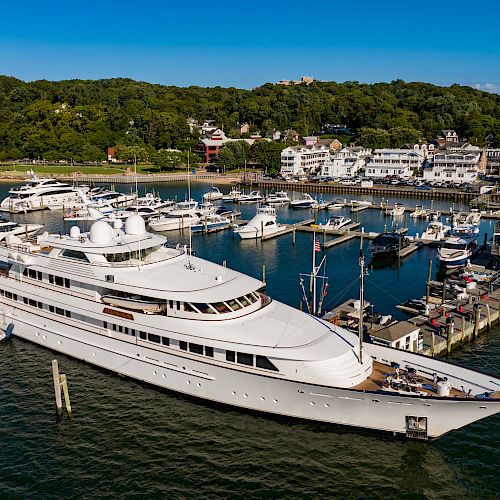 The image shows a luxurious white yacht docked in a marina, surrounded by other boats, with a scenic backdrop of greenery and houses.