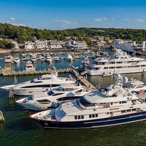 A marina filled with yachts and smaller boats docked, with a backdrop of coastal buildings and lush green hills under a clear blue sky.