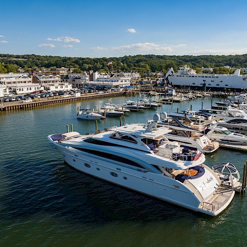 A marina filled with various boats and yachts docked at piers, with a coastal town and a large ferry in the background, under a clear blue sky.