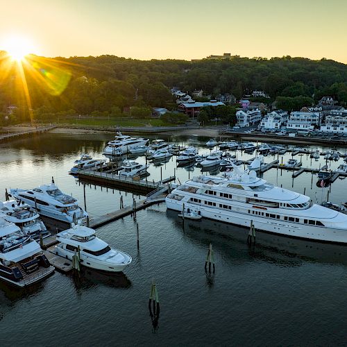 The image depicts a marina with several yachts docked, against the backdrop of a setting sun and surrounding hills and buildings.