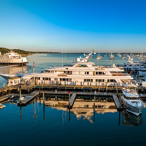 A marina filled with various yachts and boats, with a ferry approaching; industrial smokestacks visible in the distance and calm water reflecting the scene.