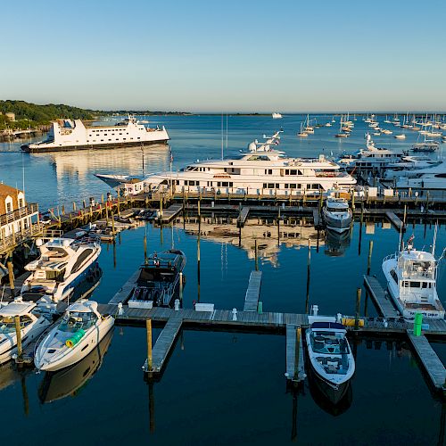 A marina with numerous yachts and boats docked, a white ferry in the background, and industrial structures on the horizon during a calm day.