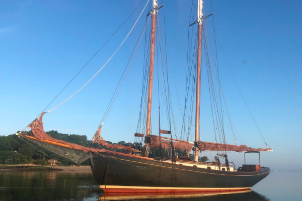 A wooden sailboat with two masts is anchored on calm water, reflecting the boat and the clear blue sky above, with a shoreline visible in the background.