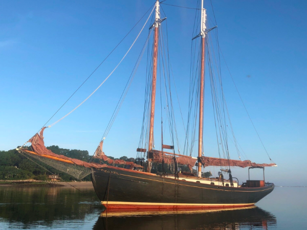 A wooden sailboat with two masts is anchored on calm water, reflecting the boat and the clear blue sky above, with a shoreline visible in the background.