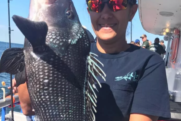 A person wearing sunglasses and a pink cap is holding a large fish on a boat, with other people and fishing rods in the background.