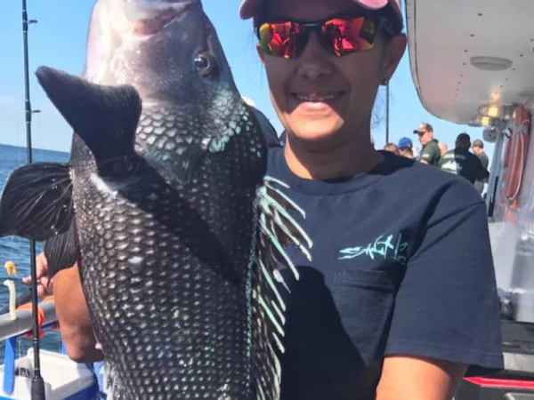 A person wearing sunglasses and a pink cap is holding a large fish on a boat, with other people and fishing rods in the background.