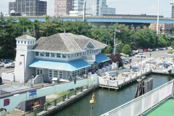 The image shows a waterfront restaurant or building near a small marina, with boats docked and a cityscape in the background.