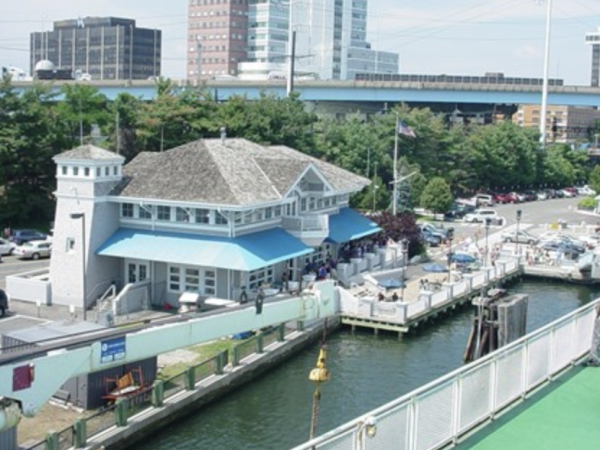 The image shows a waterfront restaurant or building near a small marina, with boats docked and a cityscape in the background.