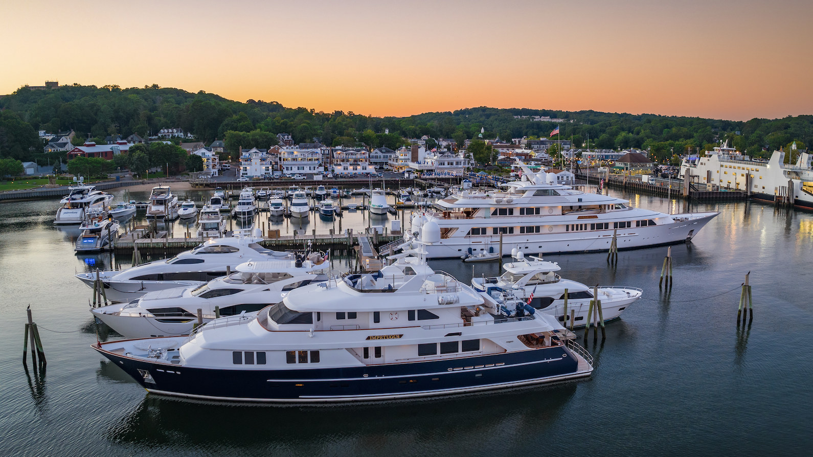 The image shows a marina filled with docks and yachts bathed in a warm sunset glow, with buildings and trees in the background filling the horizon.